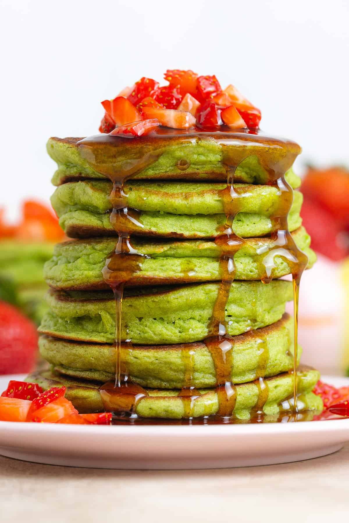 A stack of green pancakes topped with chopped strawberries and drizzled with maple syrup on a small plate.