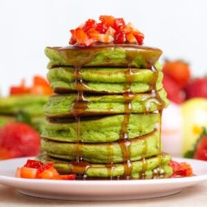 A stack of green pancakes topped with chopped strawberries and drizzled with maple syrup on a small plate.