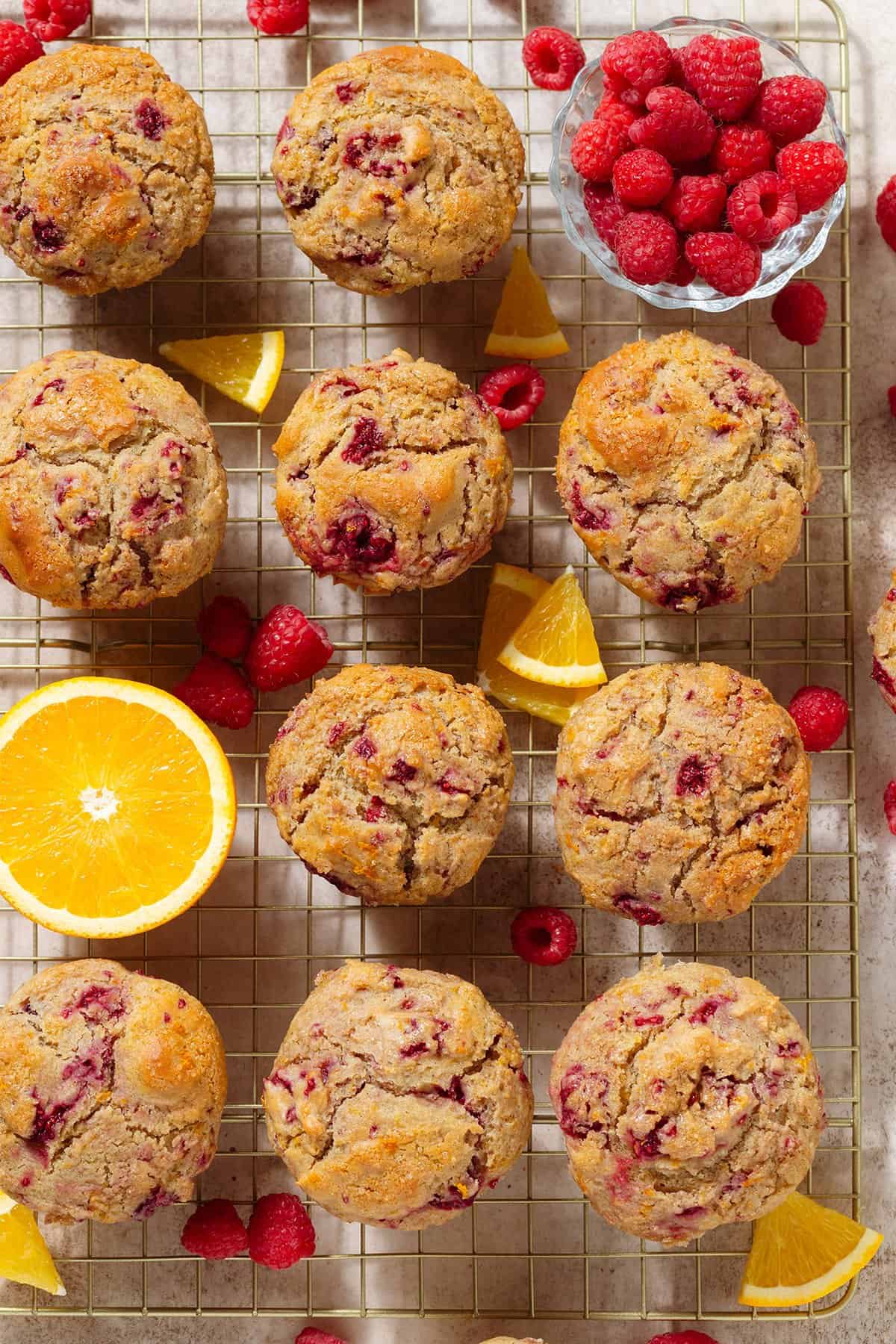 Raspberry orange muffins on a golden cooling rack with fresh raspberries and slices of orange around the muffins.