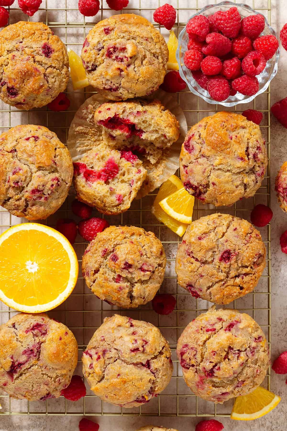 Raspberry orange muffins on a golden cooling rack with one muffin torn in half to show the inside and fresh raspberries and slices of orange around the muffins.