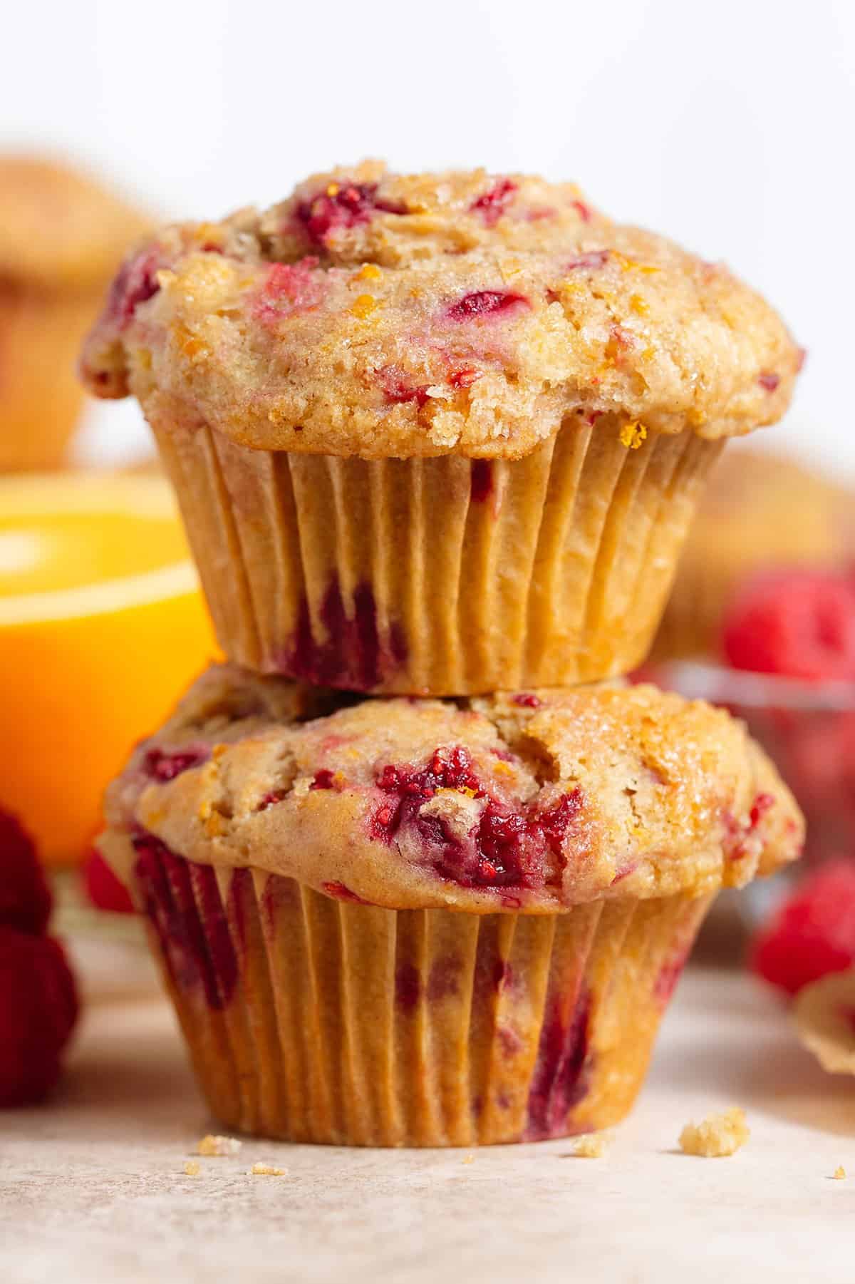 Two raspberry muffins stacked on top of each other with more muffins, raspberries, and an orange around.