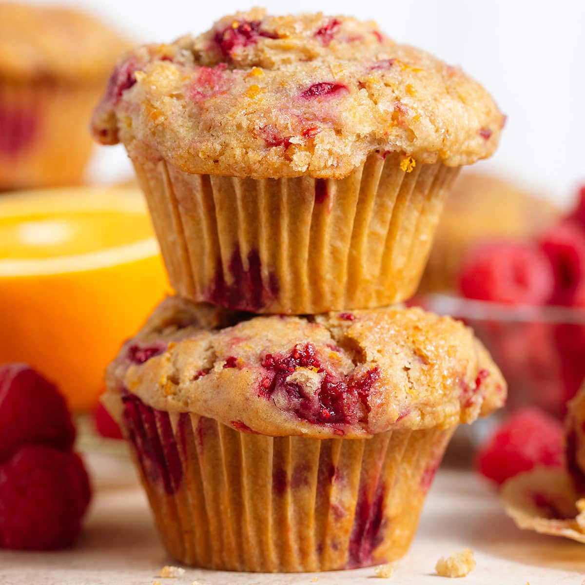 Two raspberry muffins stacked on top of each other with more muffins and raspberries around.