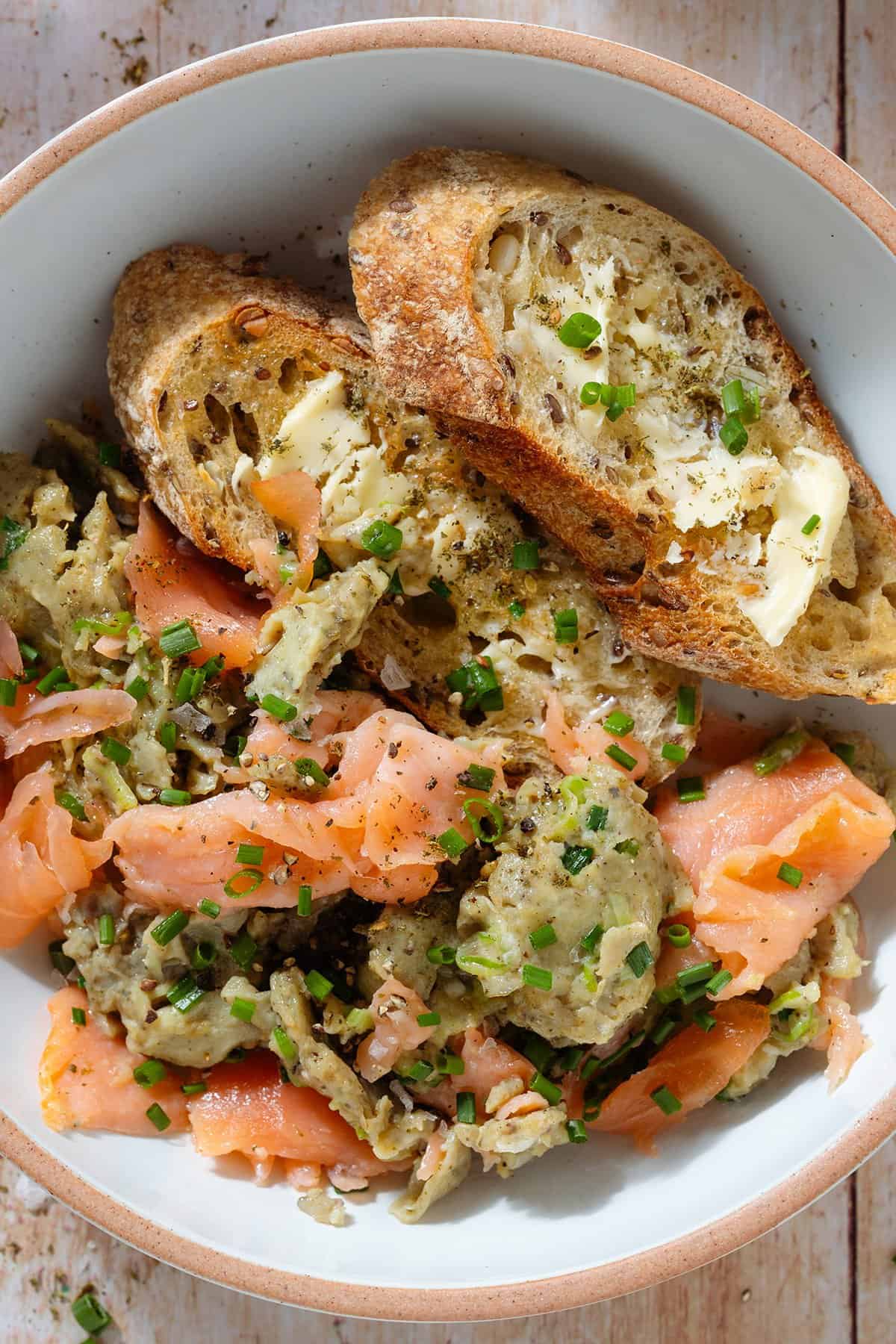 Scrambled eggs with smoked salmon in a white low bowl with toasted and buttered thick baguette slices and a fork.