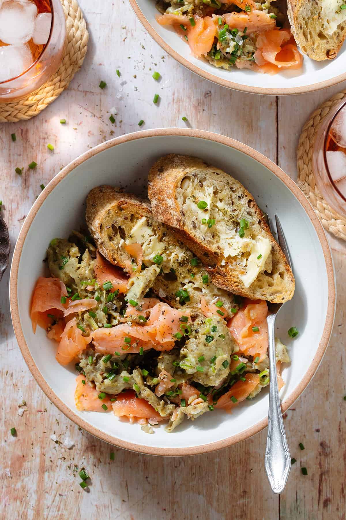 Scrambled eggs with smoked salmon in a white low bowl with toasted and buttered thick slices of baguette and a fork.