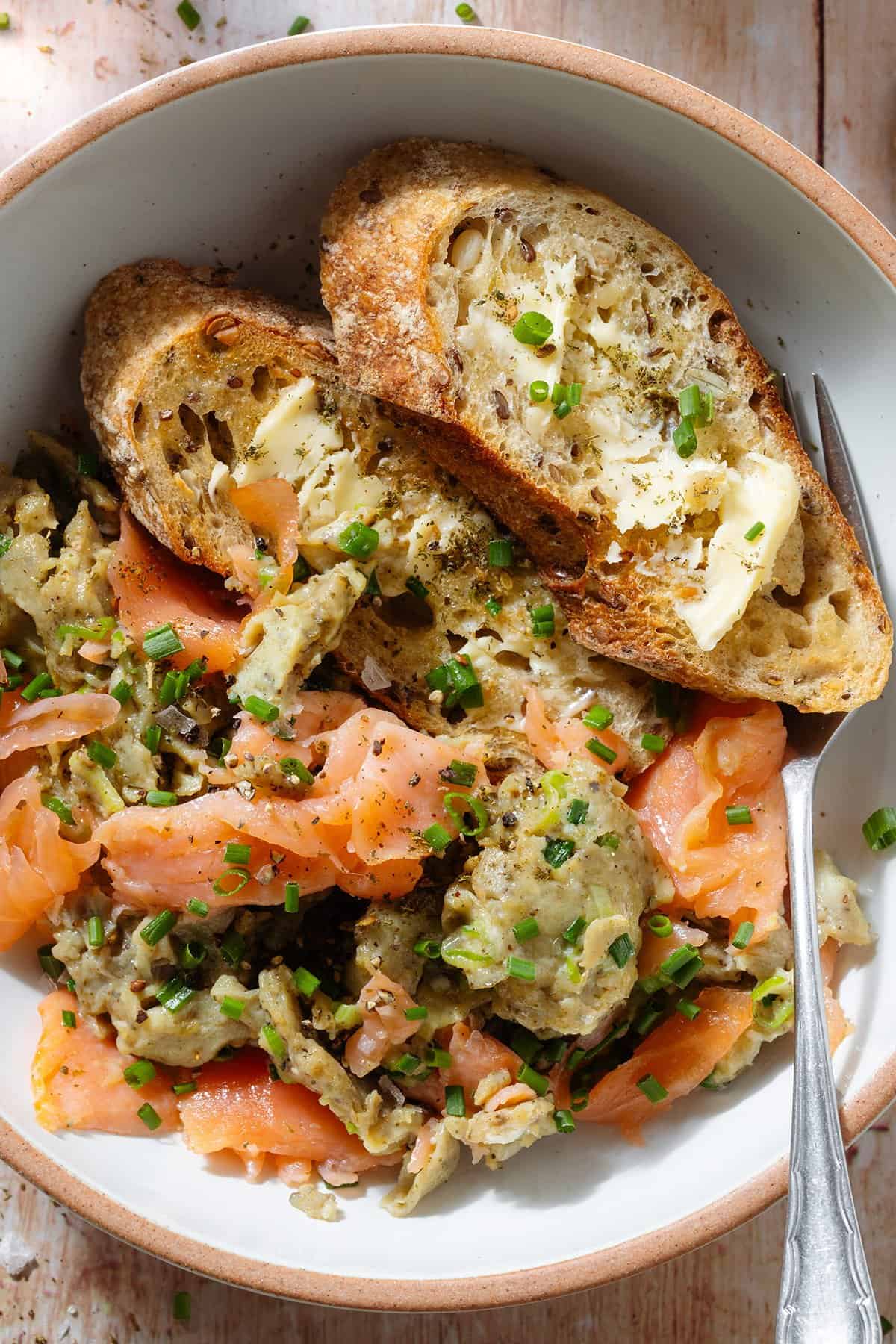 Scrambled eggs with smoked salmon in a white low bowl with toasted and buttered slices of baguette and a fork.