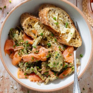 Scrambled eggs with smoked salmon in a white low bowl with toasted and buttered thick baguette slices and a fork.