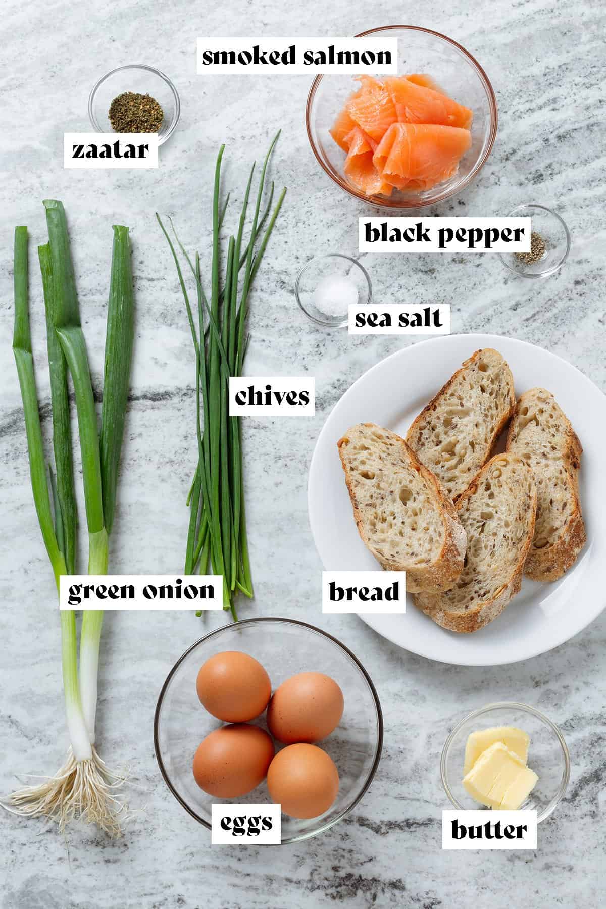 Ingredients like bread, eggs, chives, green onion, and smoked salmon on a grey stone background.