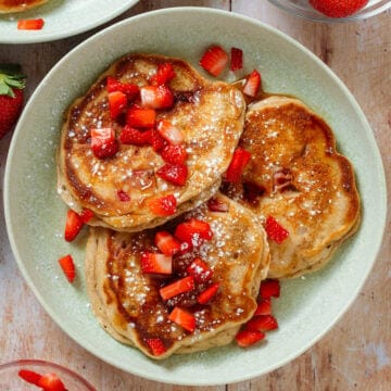Pancakes topped with fresh strawberries, maple syrup, and powdered sugar on a light green plate.