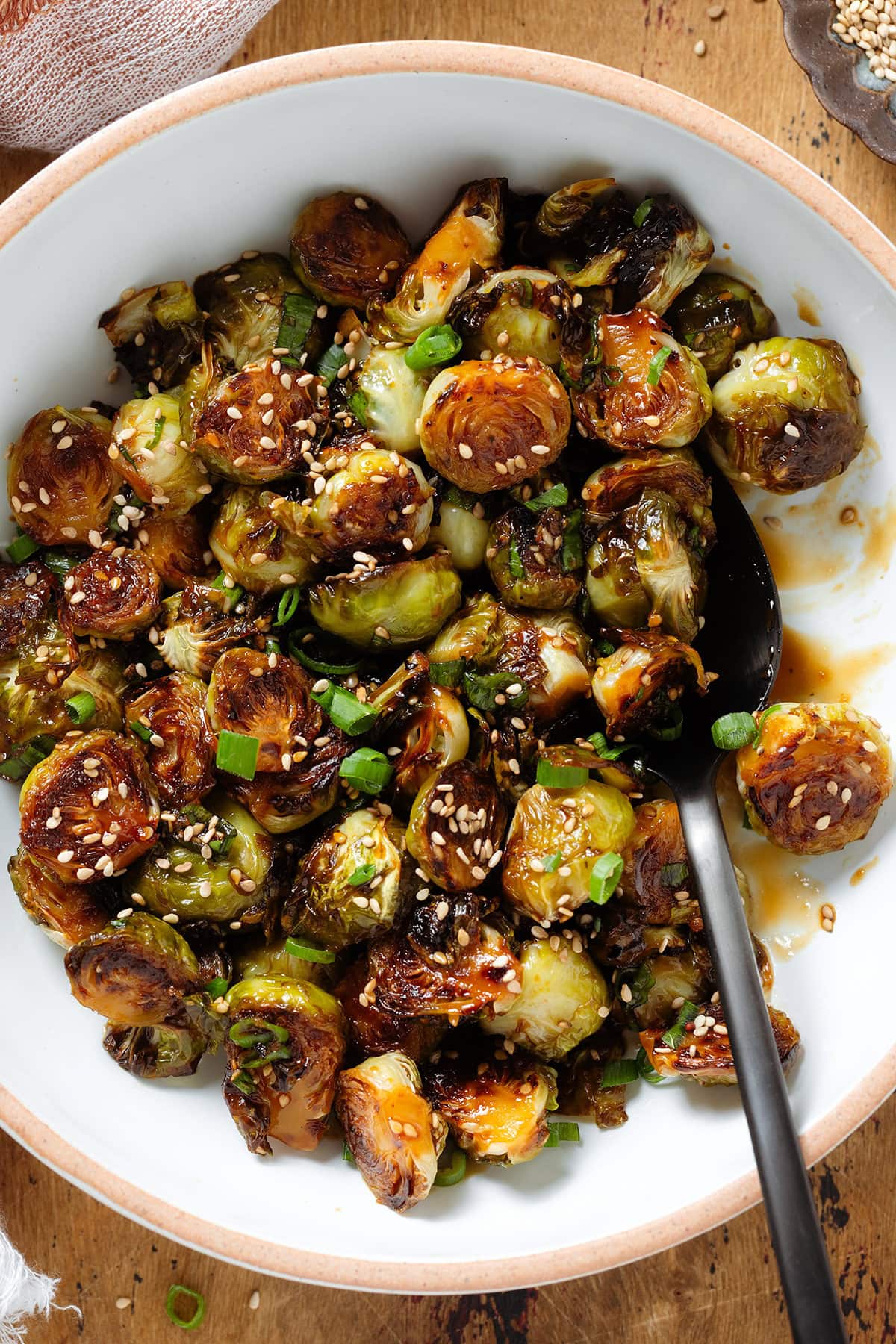 Roasted brussels sprouts sprinkled with sesame seeds and green onion in a white bowl with a beige rim and with a black spoon in the bowl.