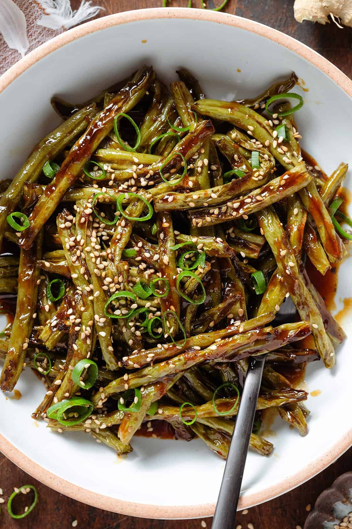 Roasted green beans with teriyaki sauce in a white bowl with a beige rim garnished with green onion and sesame seeds.