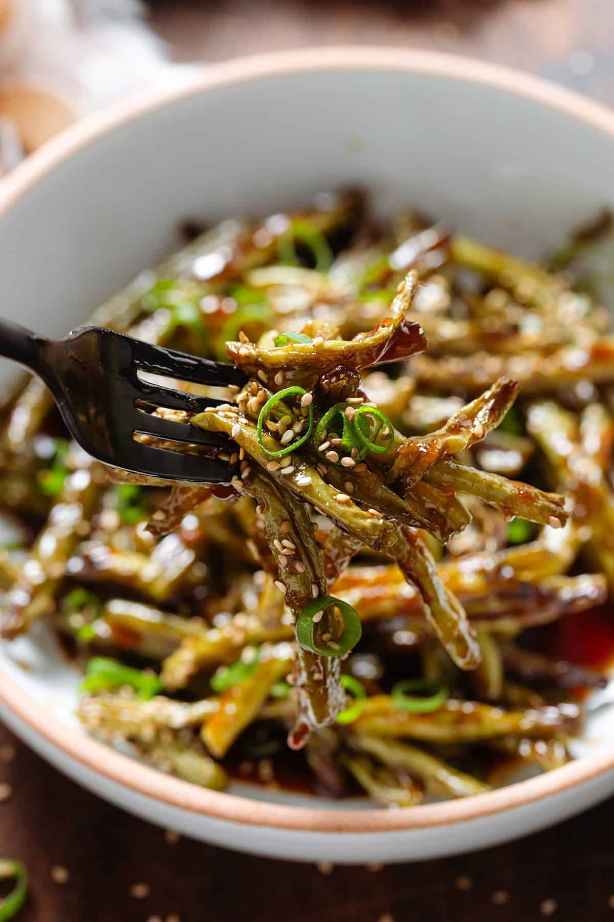 A fork holding roasted green beans with golden teriyaki sauce garnished with green onion and sesame seeds.