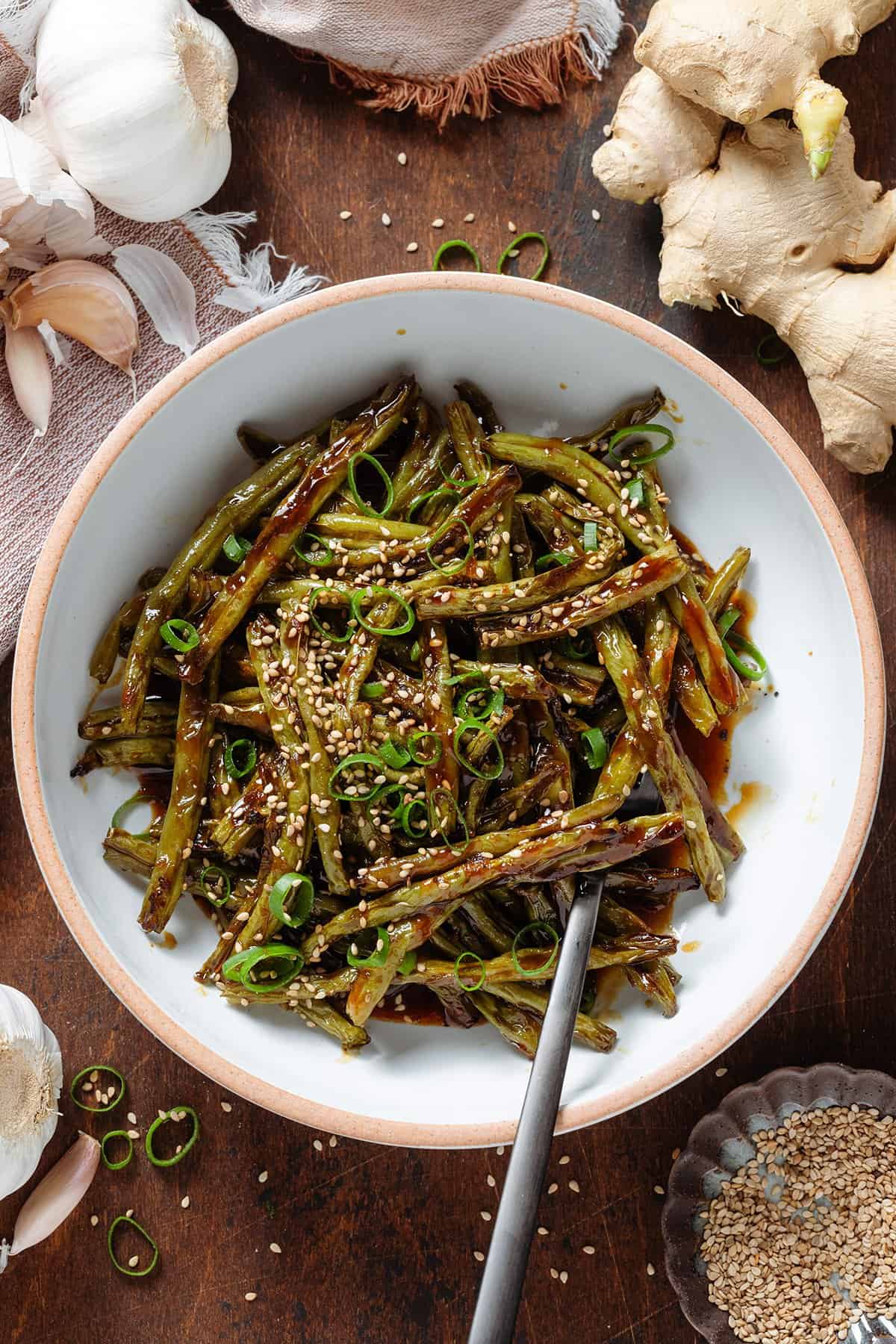 Roasted green beans with teriyaki sauce in a white bowl with a beige rim garnished with green onion and sesame seeds.