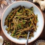 Roasted green beans with teriyaki sauce in a white bowl with a beige rim garnished with green onion and sesame seeds.
