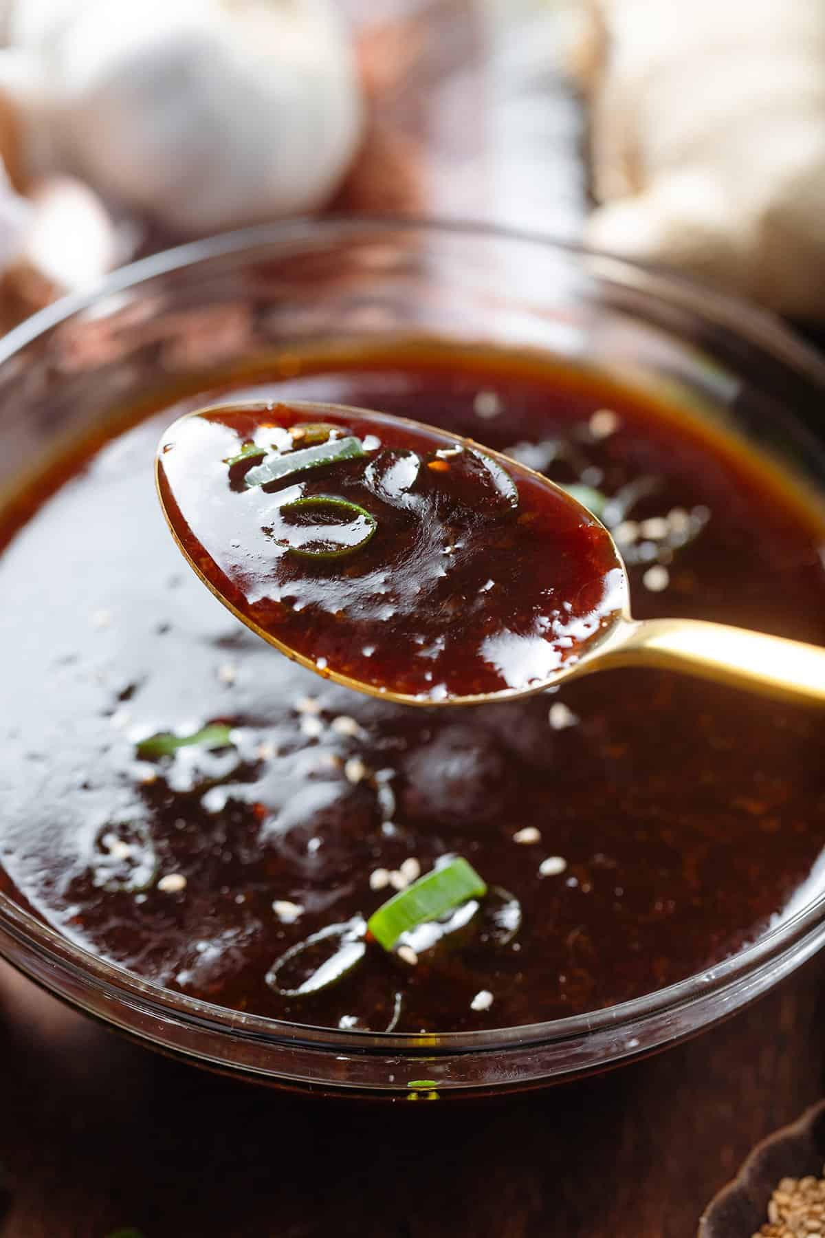 A spoon holding dark teriyaki sauce over a glass bowl with more of the sauce garnished with green onion and sesame seeds.