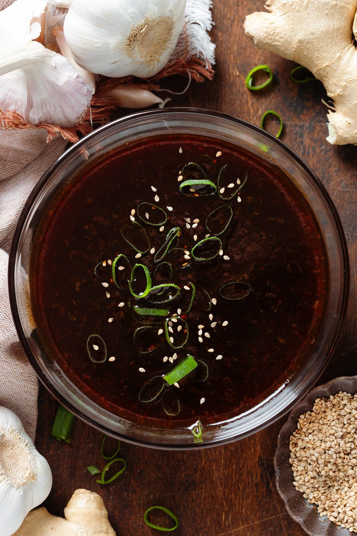 Dark teriyaki sauce in a glass bowl garnished with green onion and sesame seeds with garlic, ginger, and more sesame seeds around the bowl.