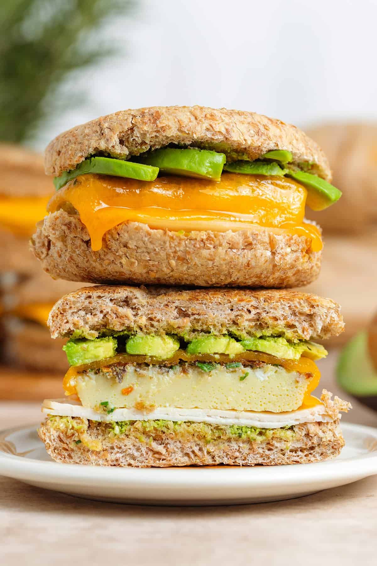 Two breakfast sandwiches with eggs, ham, melted cheese, and avocado stacked on top of each other, the bottom one cut in half showing the inside.