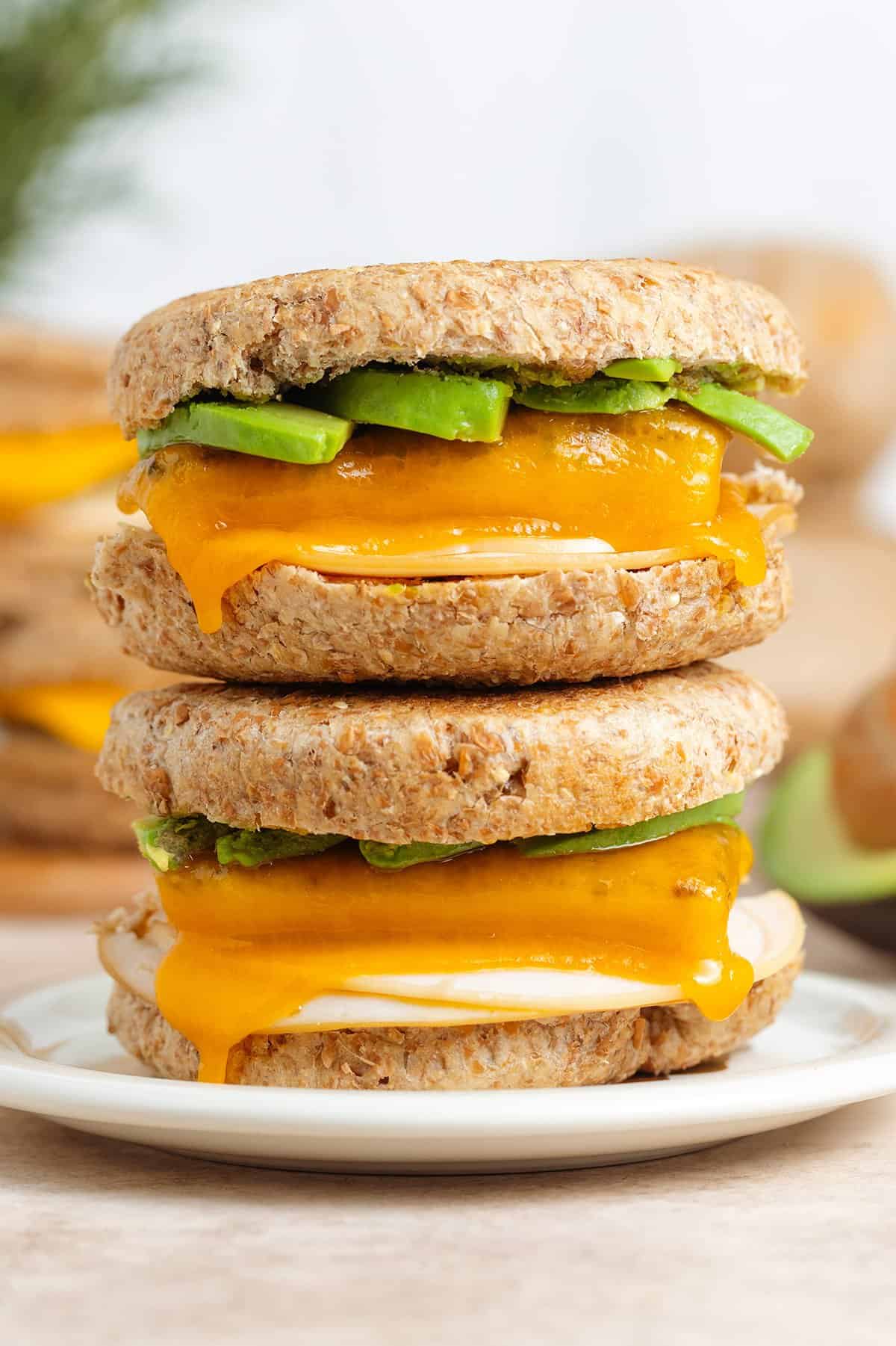 Two breakfast sandwiches with ham, melted cheese, and avocado stacked on top of each other on a small plate.
