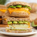 Two breakfast sandwiches with eggs, ham, melted cheese, and avocado stacked on top of each other, the bottom one cut in half showing the inside.