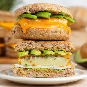 Two breakfast sandwiches with eggs, ham, melted cheese, and avocado stacked on top of each other, the bottom one cut in half showing the inside.