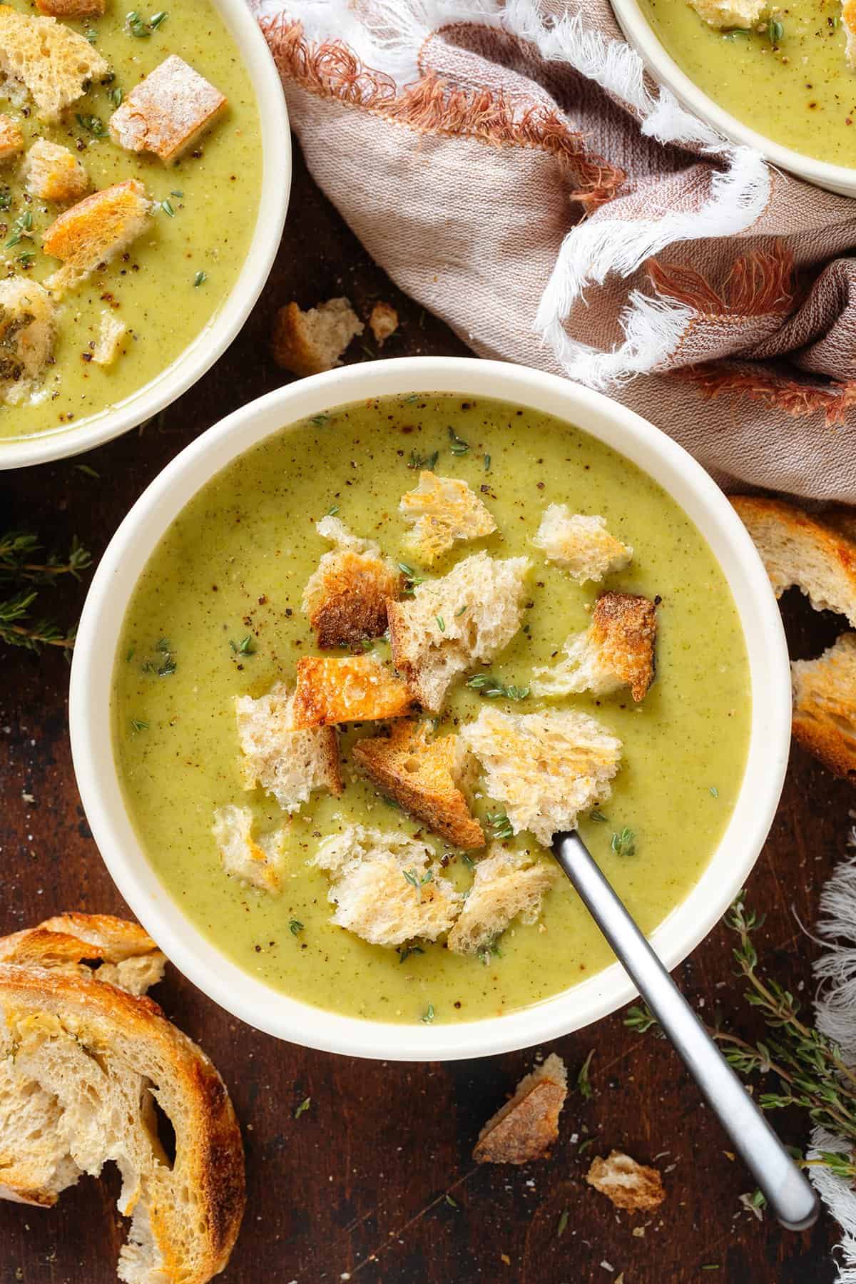 Creamy green broccoli soup in white bowls garnished with torn up toasted sourdough bread and fresh thyme with a black spoon in the bowl in the middle.