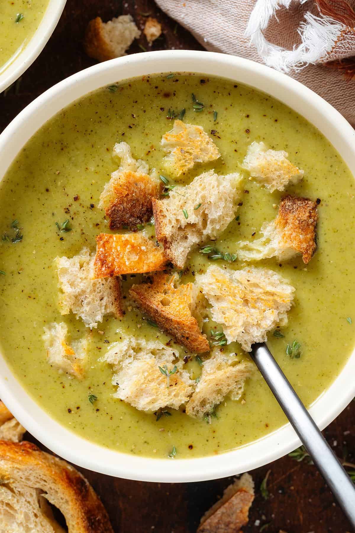 Creamy green broccoli soup in a white bowl garnished with torn up toasted sourdough bread and fresh thyme.