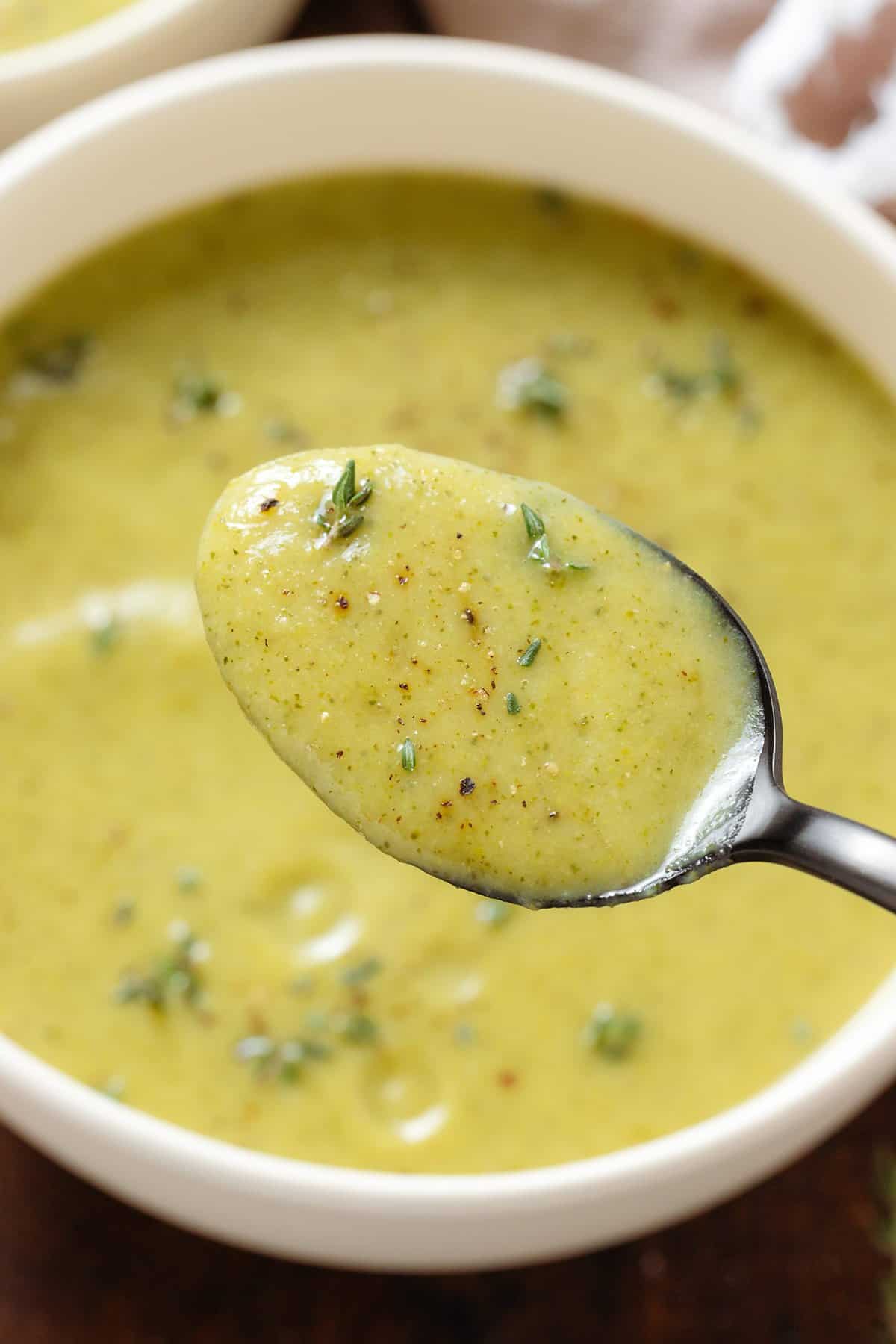 A black spoon holding creamy broccoli zucchini soup with fresh thyme over a bowl of the soup.