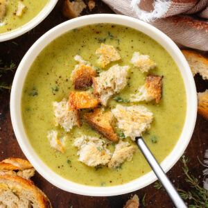 Creamy green broccoli soup in a white bowl garnished with torn up toasted sourdough bread and fresh thyme.