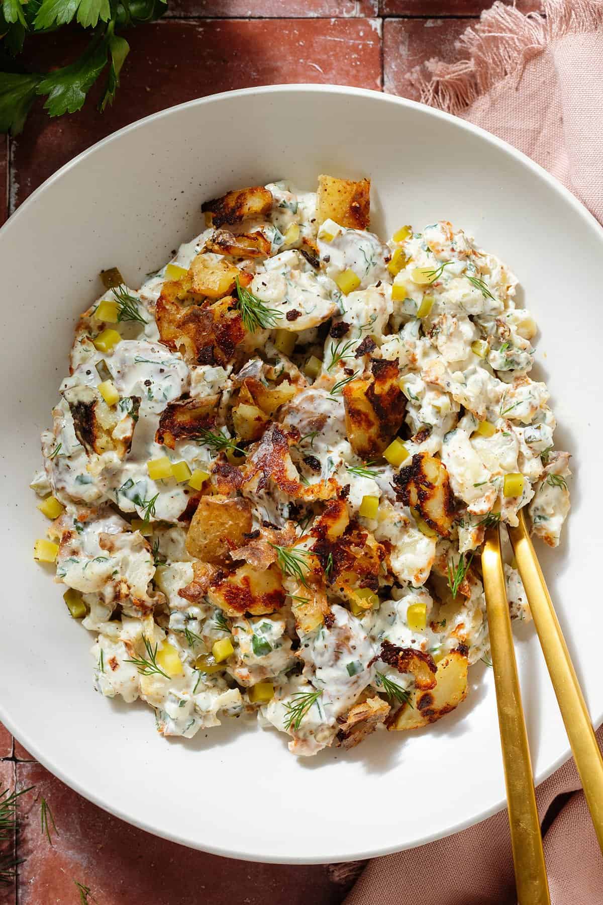 Crispy potato salad with yogurt mayo dressing garnished with bit of more crispy potatoes in a white low bowl with gold serving spoons.