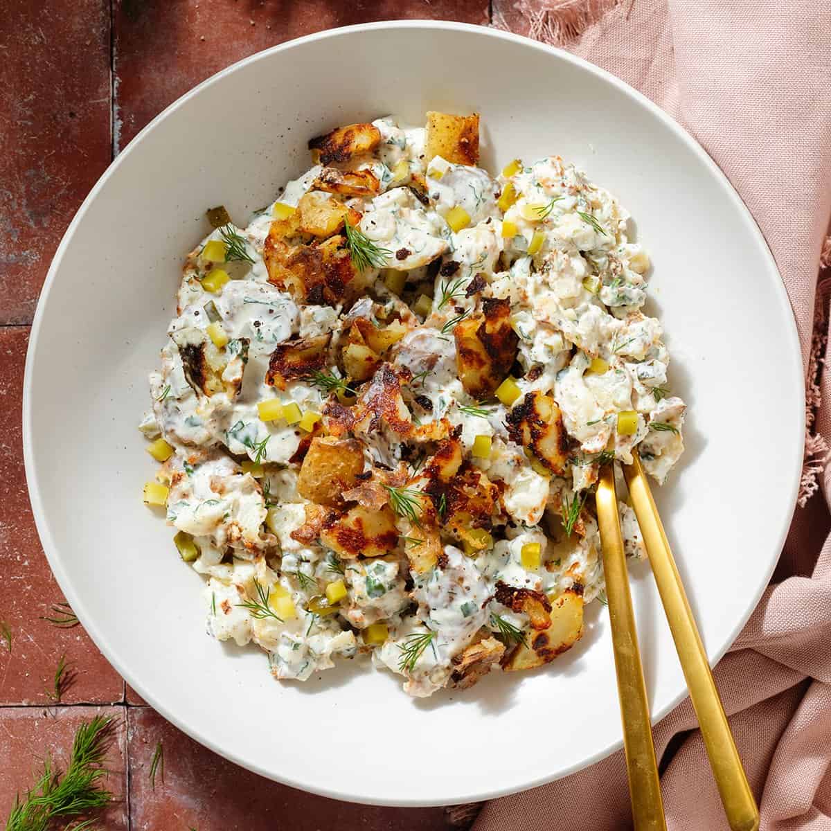 Crispy potato salad with yogurt mayo dressing garnished with bit of more crispy potatoes and fresh dill in a white bowl.