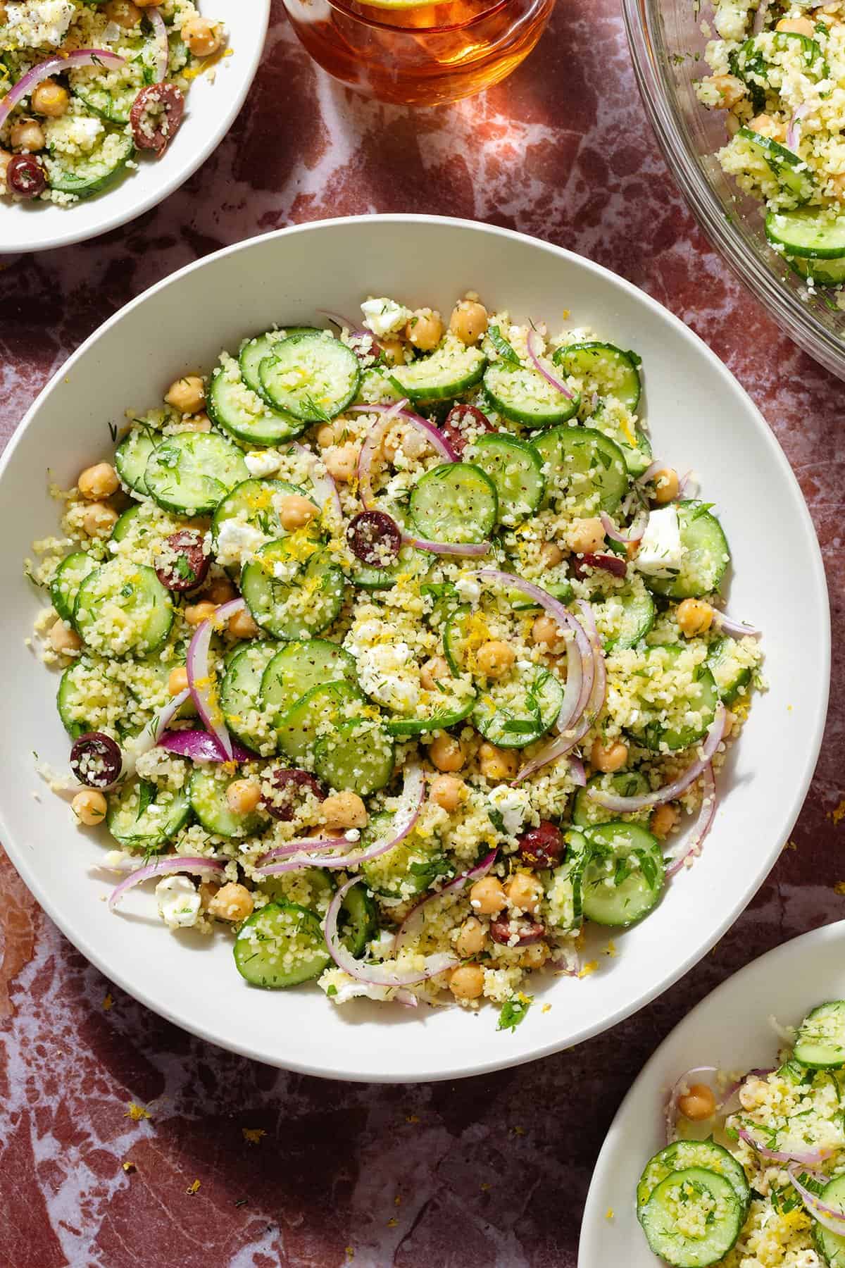 Cucumber and couscous salad in white bowls with gold utensils on a marble background.
