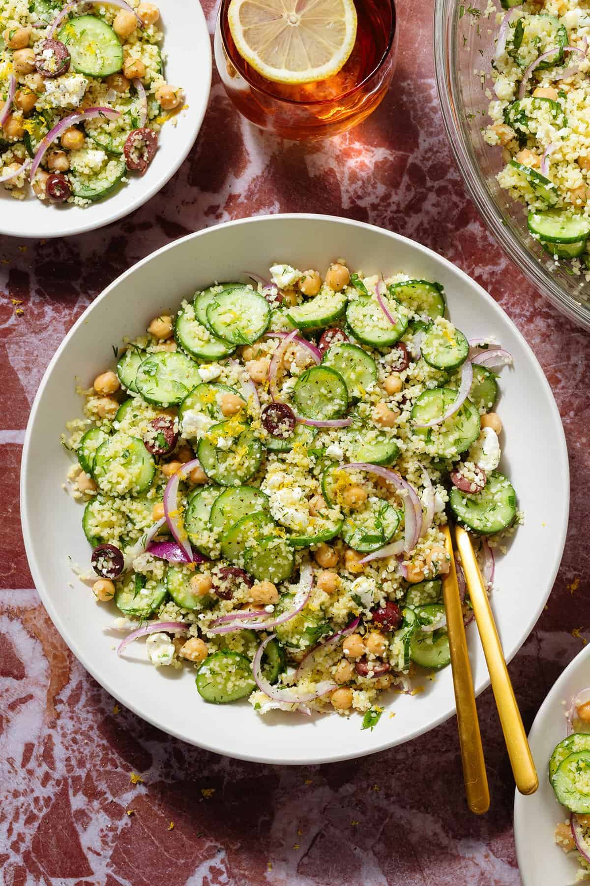Cucumber and couscous salad in white bowls with gold utensils on a marble background.