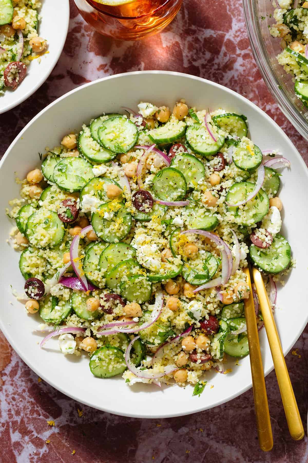 Cucumber and couscous salad in a large white bowl with gold utensils on a marble background.