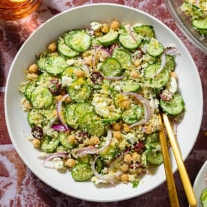 Cucumber and couscous salad in a large white bowl with gold utensils on a marble background.