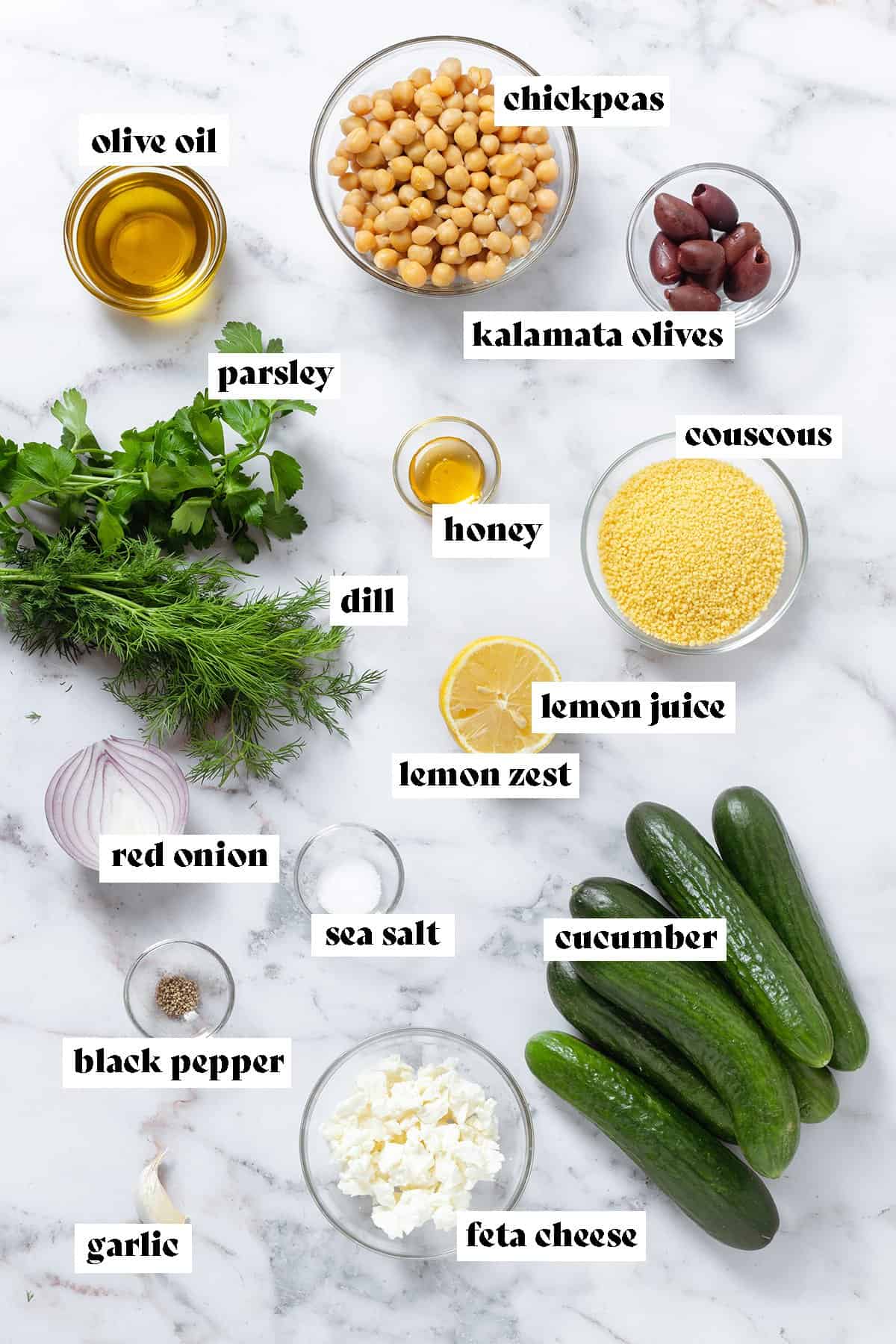 Fresh herbs, mini cucumbers, chickpeas, olives, feta, and other ingredients measured and laid out on a marble background.