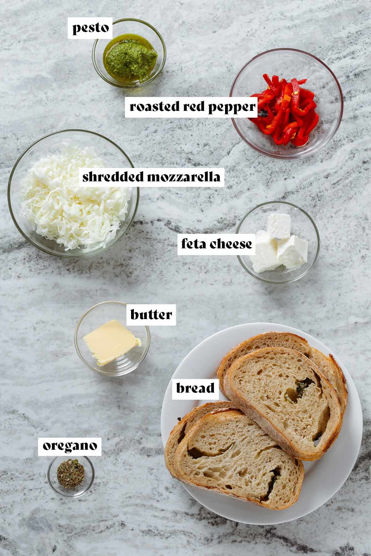 Ingredients like sliced bread, shredded mozzarella, sliced red peppers, feta cheese, pesto, and spices in small bowls all laid out.