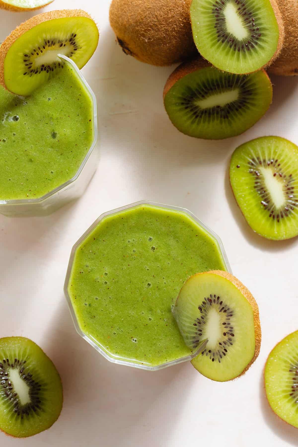 Bright green smoothie in two short glasses garnished with kiwi slices and more kiwi fruit around.