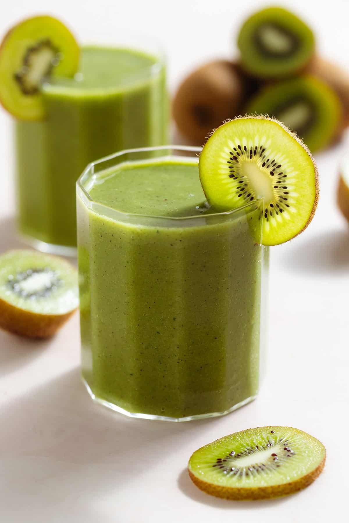 Bright green smoothie in two short glasses garnished with kiwi slices and more kiwi fruit around.