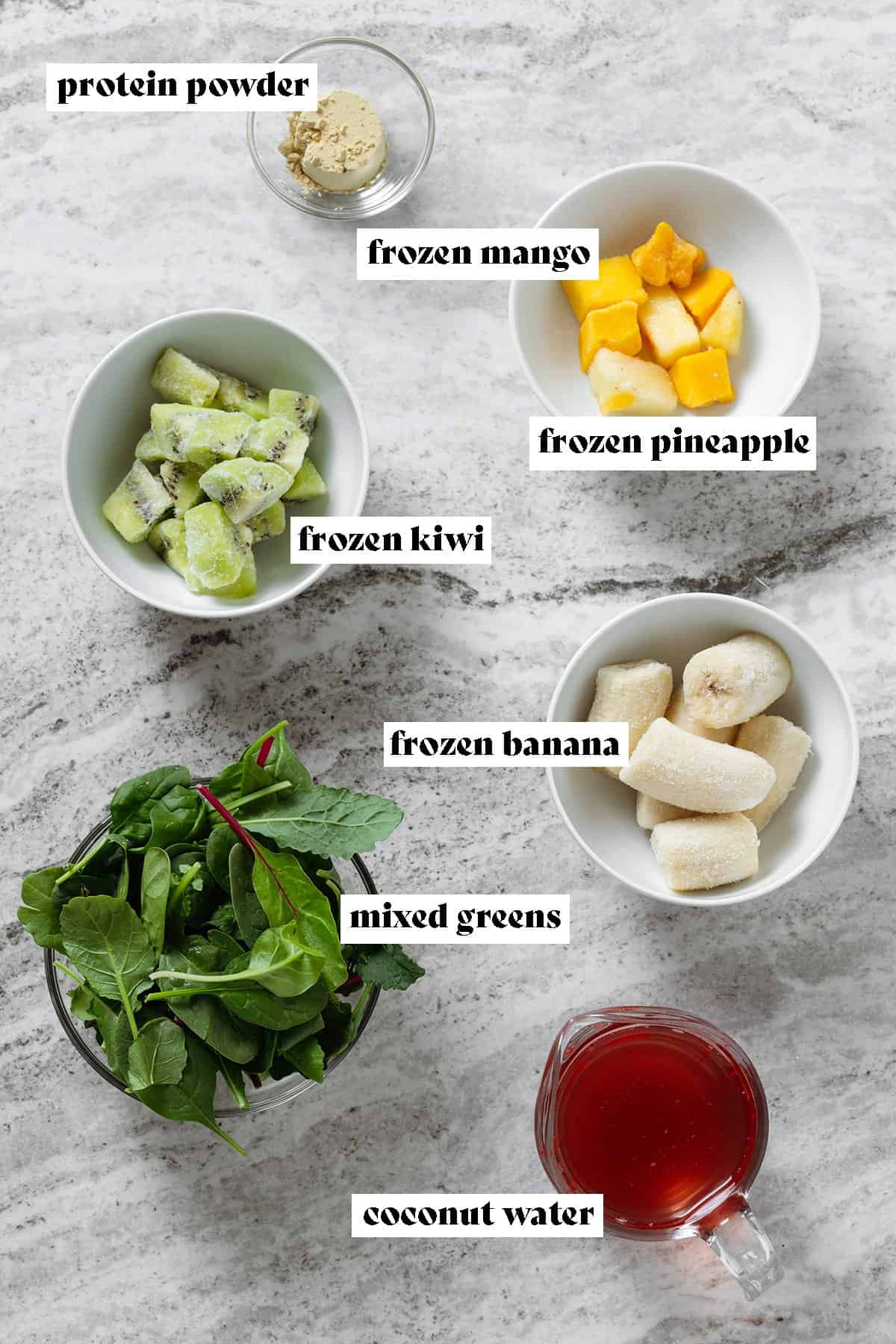 Small bowls with frozen kiwi, banana, mango, mixed greens, and coconut water on a stone background.