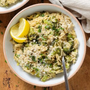 Creamy broccoli orzo in a white bowl with a beige rim garnished with two lemon wedges and sprinkled with parmesan.