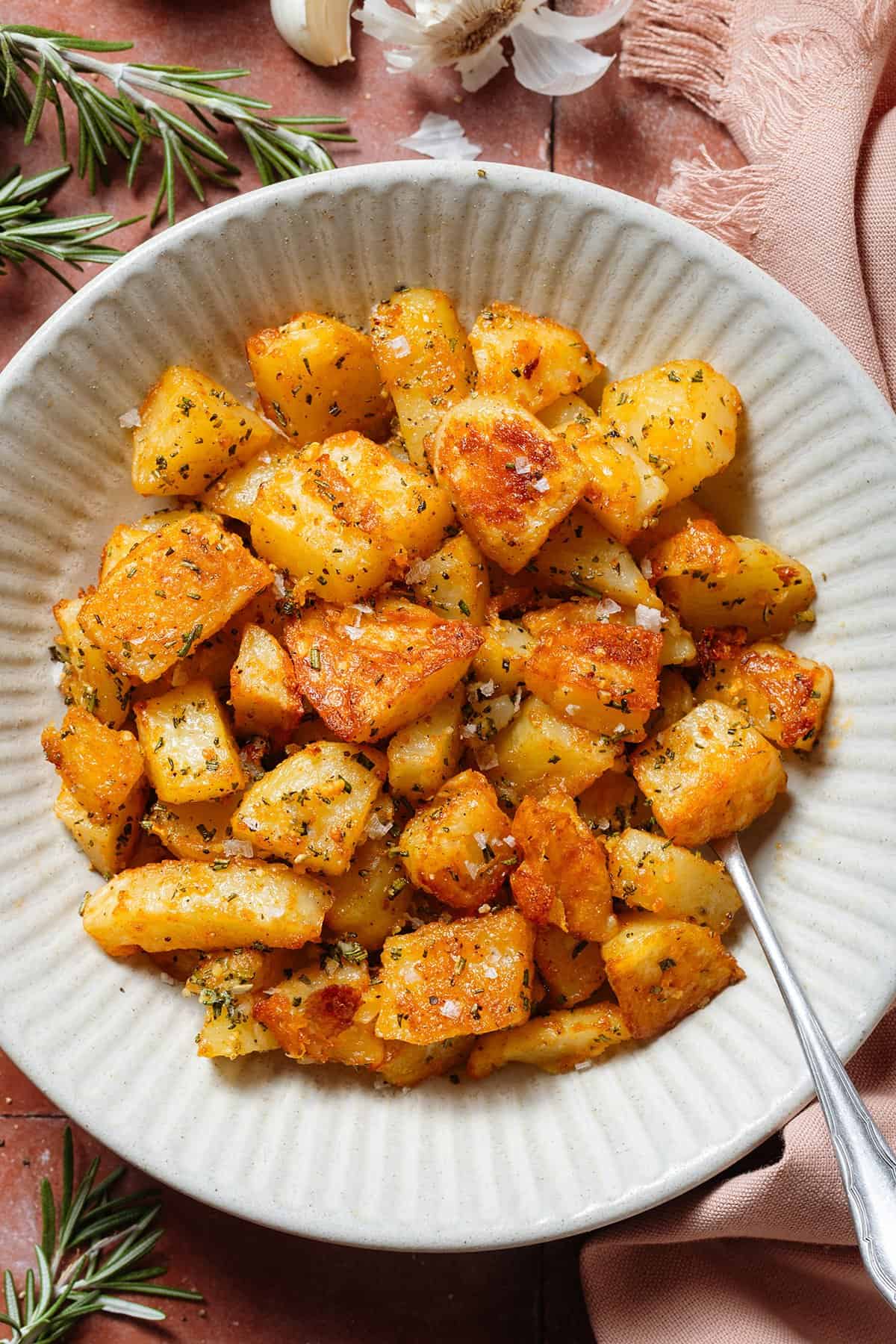 Crispy roasted potatoes with specks of chopped rosemary, minced garlic, and flaky sea salt in a white bowl.