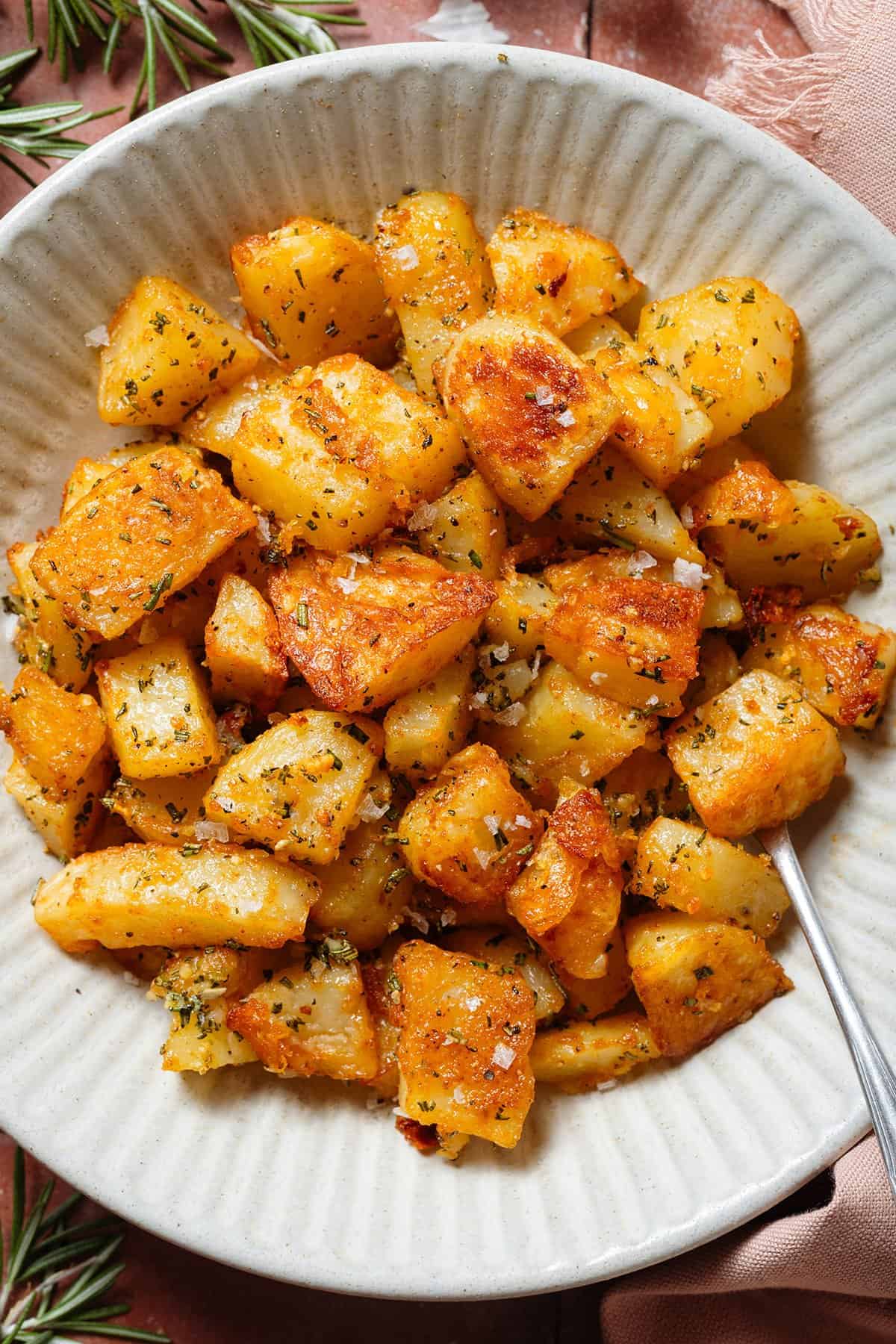 Crispy roasted potatoes with specks of chopped rosemary, minced garlic, and flaky sea salt in a white bowl.