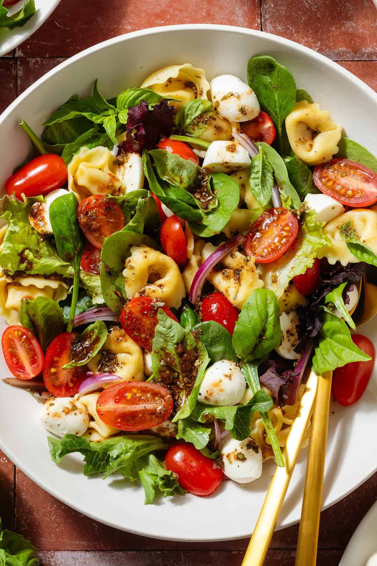 Tortellini caprese salad with mixed greens drizzled with balsamic dressing in a large white bowl with gold serving spoons.