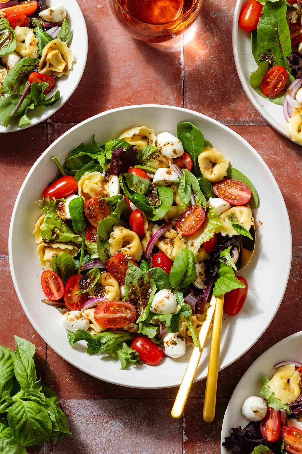Tortellini caprese salad with mixed greens in a white bowls with gold serving spoons in the one in the middle.