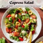 Tortellini caprese salad with mixed greens in a white bowl with gold serving spoons.