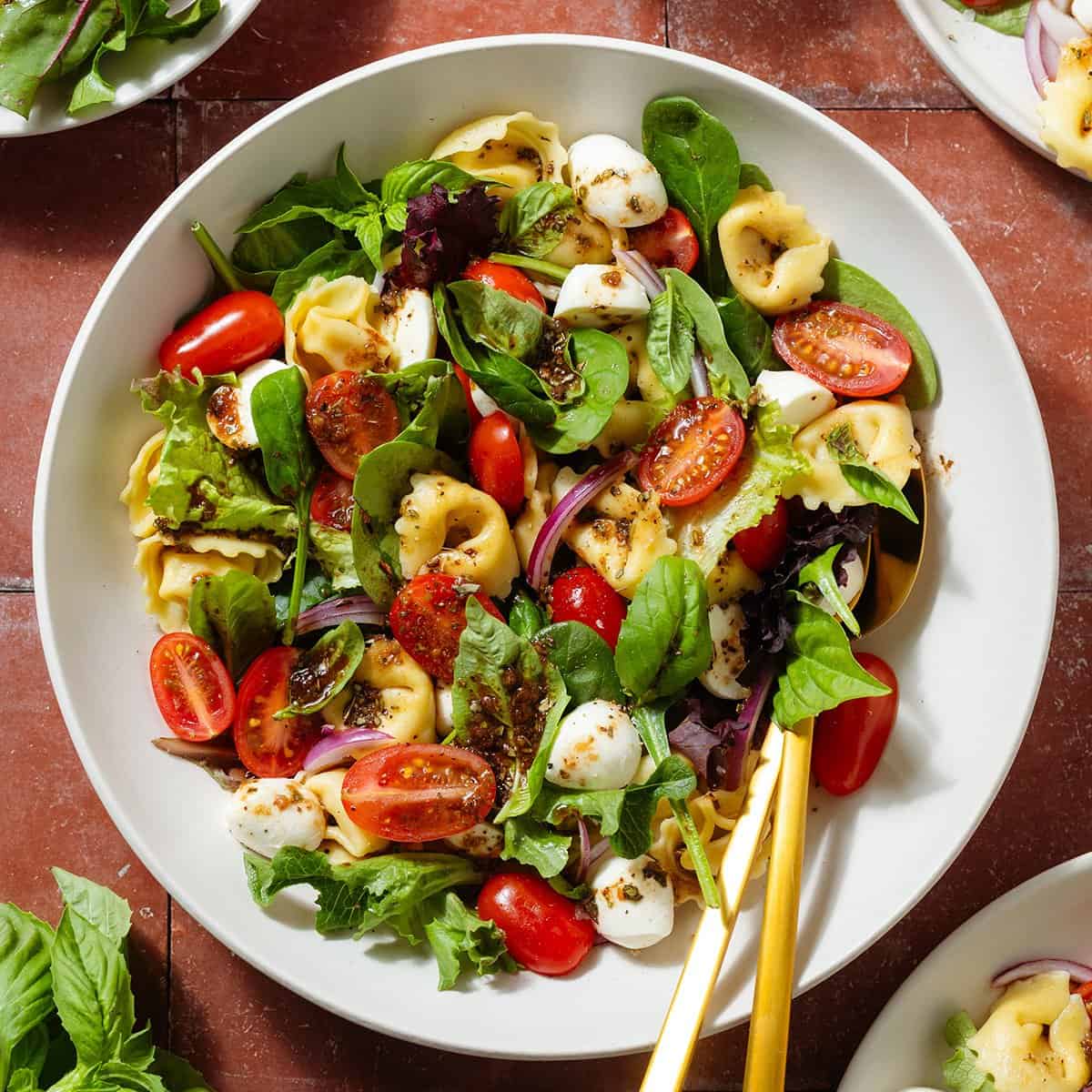 Tortellini caprese salad with mixed greens in a white bowl with gold serving spoons.