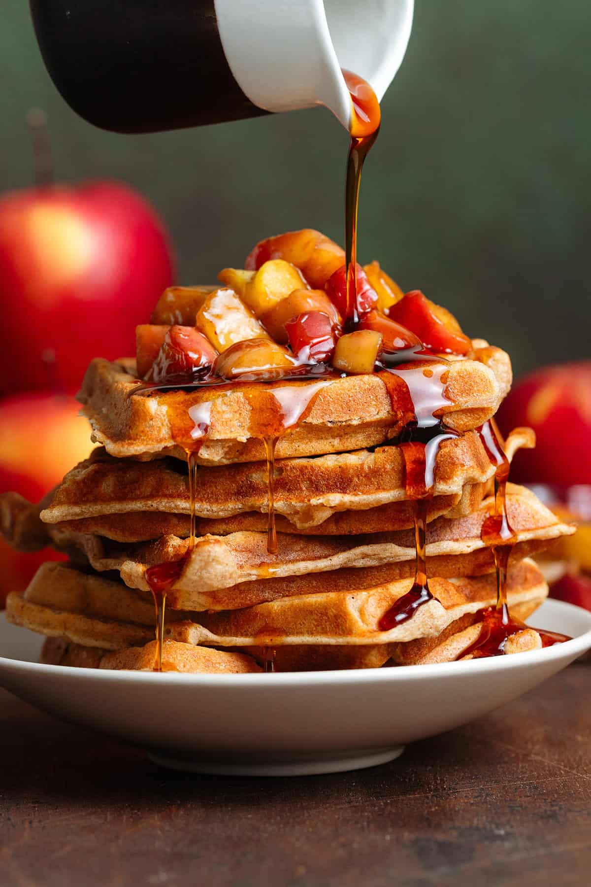 A stack of waffles with caramelized apples on top and maple syrup being drizzled over them.