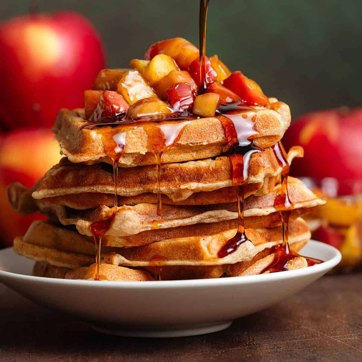 A stack of waffles with caramelized apples on top and maple syrup being drizzled over them.