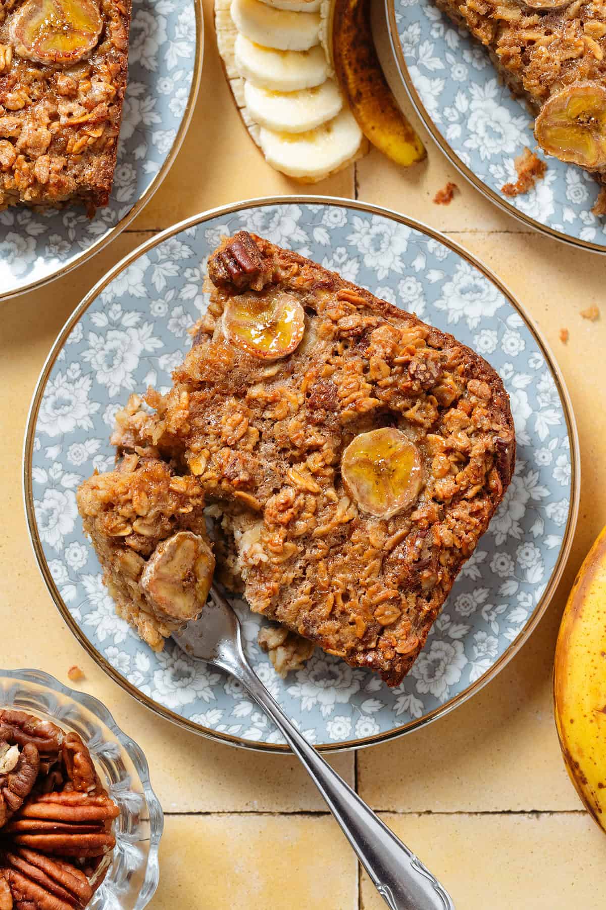 A slice of banana baked oatmeal with caramelized banana slices baked into it on top on blue floral plates with a fork holding a bite of the corner of the slice.