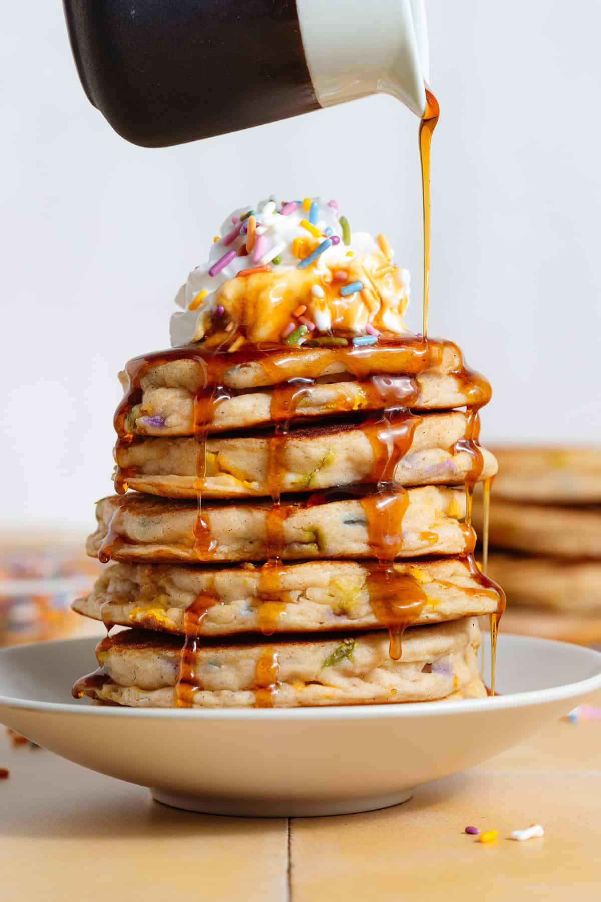 A stack of pancakes with sprinkles inside and whipped cream on top being drizzled with maple syrup.