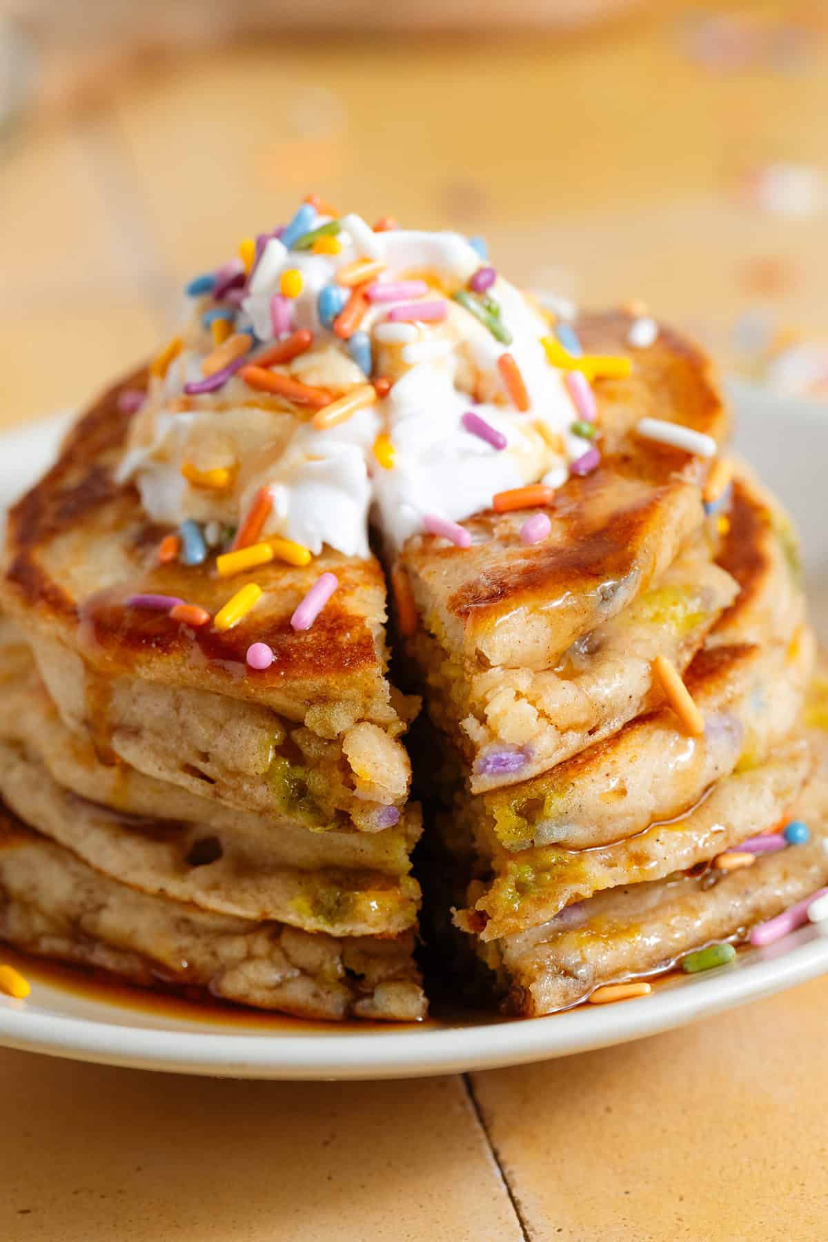 A small stack of pancakes with sprinkles with whipped cream and maple syrup on top.